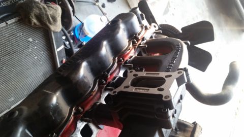 1985 Ford Bronco - A close-up view of a black engine manifold with a carburetor spacer installed. A fan is visible behind the manifold, and a radiator is part…