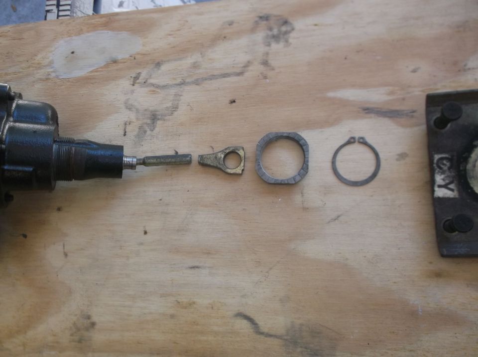 Disassembled mechanism parts on a wooden surface: black cylinder, brass fitting, metal ring, retaining clip.