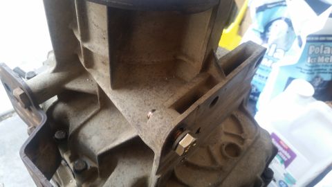 1985 Ford Bronco - This is a close-up, angled view of a rusty, cast-iron transmission housing with a metal bracket bolted to it. A hex bolt with a copper wash…