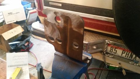 1985 Ford Bronco - A rusty metal bracket with three holes is clamped in a blue vise. In the background, parts of a vehicle, including a red stripe and a licen…
