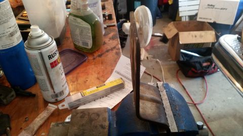 1985 Ford Bronco - A workbench scene with various items including spray cans, a vise holding a metal piece, a fan, and boxes in the background.