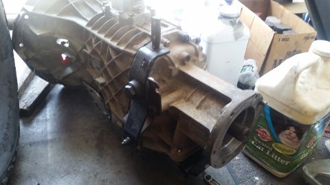 1985 Ford Bronco - A heavy-duty transmission assembly is shown in a workshop setting. The transmission is mounted on a stand, with various containers and a ti…