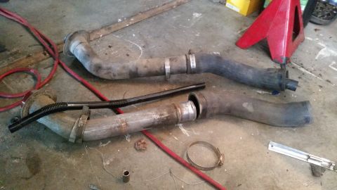 1985 Ford Bronco - Two exhaust pipes with clamps and a black flexible hose are laid out on a concrete floor next to red jumper cables and a red jack stand.