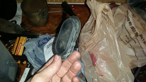 1985 Ford Bronco - A hand holds a black rubber oval-shaped part over a wooden surface with various items including plastic bags and a drill bit set.