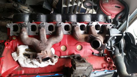 1985 Ford Bronco - A red V8 engine block with exhaust manifolds attached is shown from a top-down perspective. The engine appears to be in a workshop or garag…