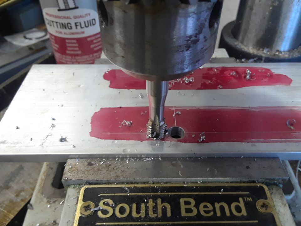 Drill bit tapping a hole in a metal plate with red markings. Cutting fluid is visible