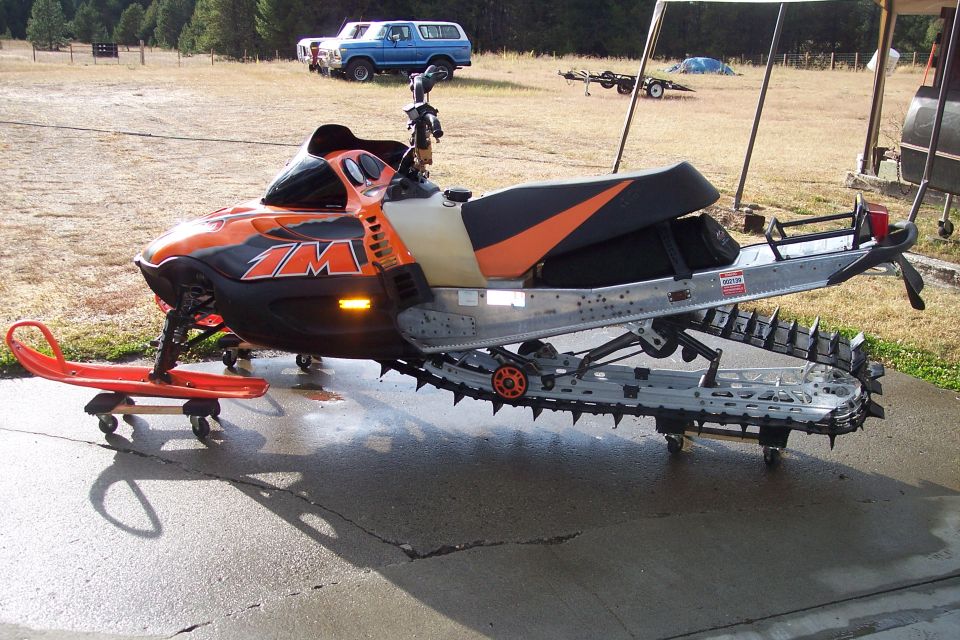 Orange and black snowmobile with a red ski on a dolly, parked on wet pavement