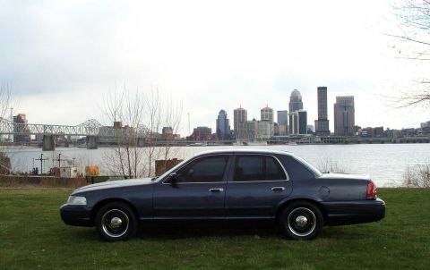 1998 Ford Crown Victoria - Side view of a 1998 Ford Crown Victoria parked on grass with a city skyline in the distance.