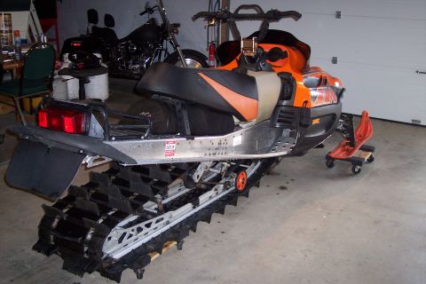 2004 Arctic Cat King Cat 900 - An orange and black snowmobile is parked indoors next to a motorcycle. The snowmobile has a large track and a red seat attachment on wheels.