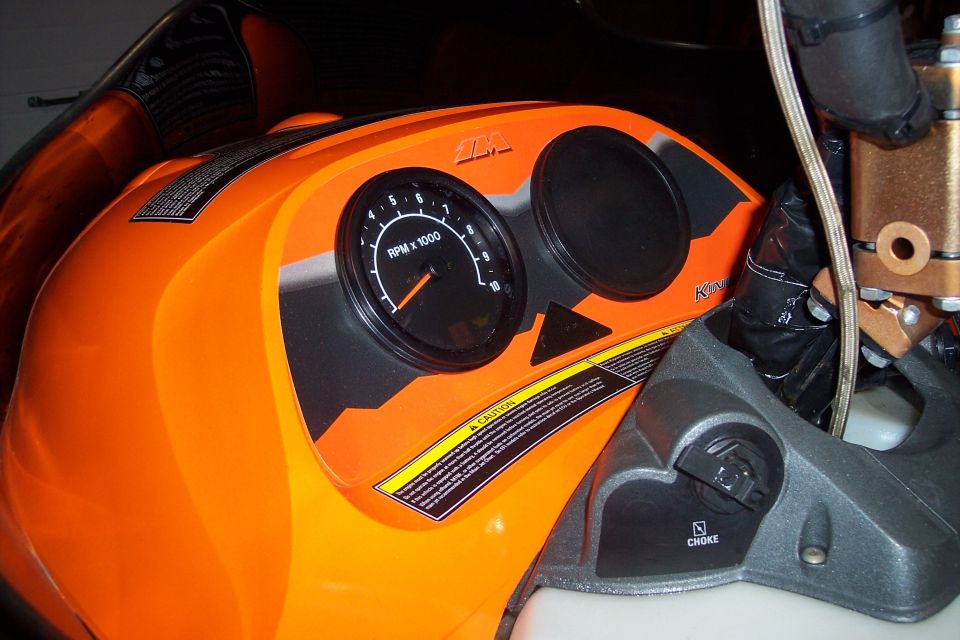 Snowmobile dash with tachometer and choke lever. Text on gauges and warning labels is visible