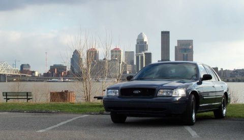 1998 Ford Crown Victoria - A dark sedan is parked near a waterfront with a city skyline visible across the water.