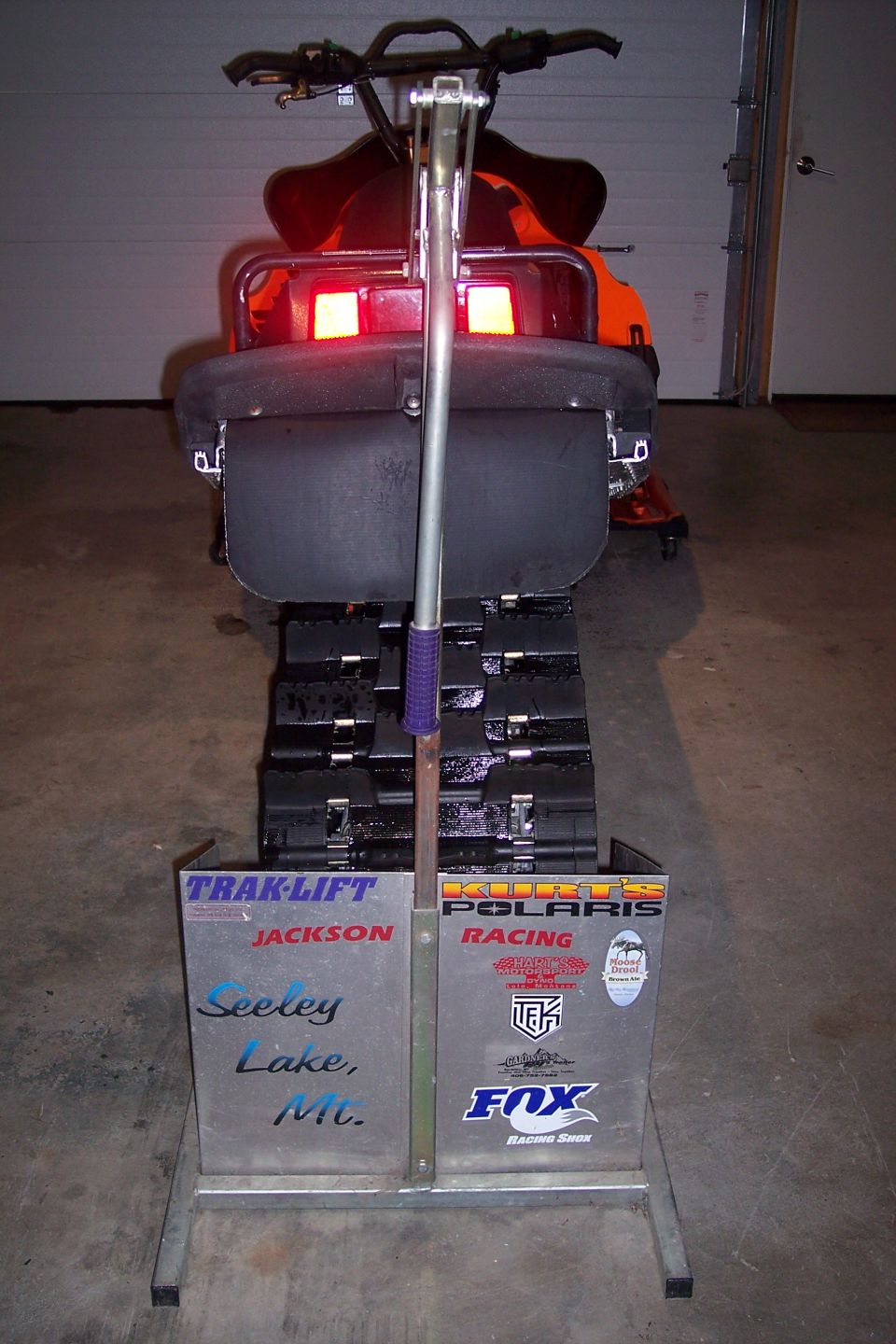 Snowmobile rear view with orange body, black seat, and red taillights. Metal plate with stickers and text is visible
