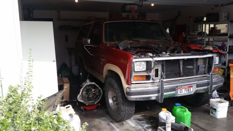 1985 Ford Bronco - A red 1985 Ford Bronco sits in a garage with its hood open and transmission removed, indicating a repair or modification is underway.