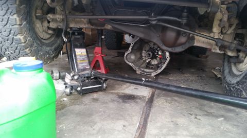 1985 Ford Bronco - Vehicle undercarriage shown with transmission removed, supported by a jack and jack stand, with a green fluid container nearby.