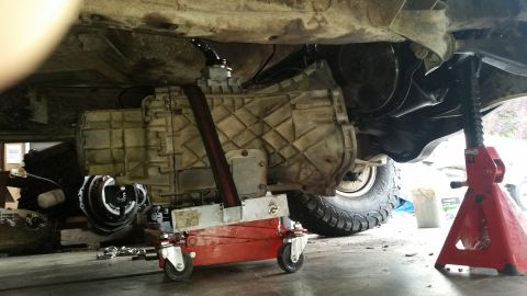 1985 Ford Bronco - Transmission being lowered from underside of vehicle with a jack, Supermotors.net logo visible.