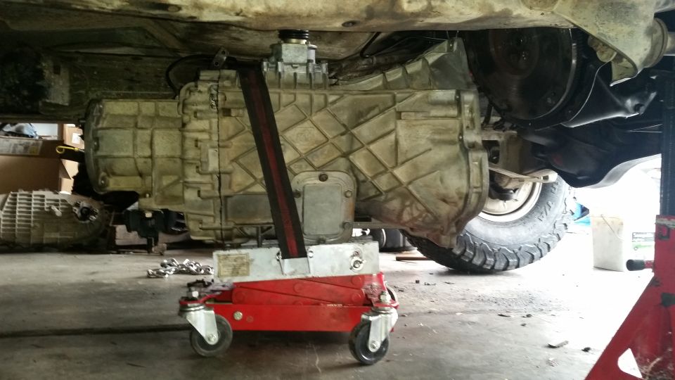 Transmission jack supporting a large transmission under a vehicle; tire and suspension visible