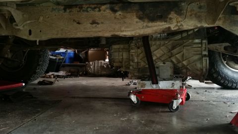 1985 Ford Bronco - Transmission being serviced under a vehicle, supported by a red hydraulic jack.