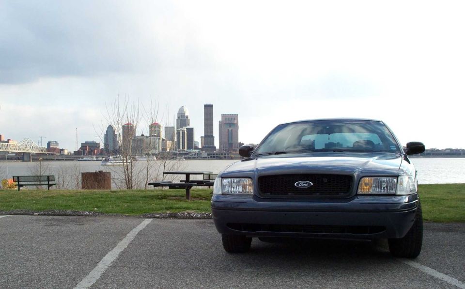 Front view of dark blue Ford Crown Victoria parked by a river with city skyline behind