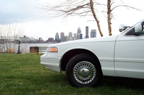 1998 Ford Crown Victoria - A white 1998 Ford Crown Victoria is parked near a waterfront with a city skyline visible.