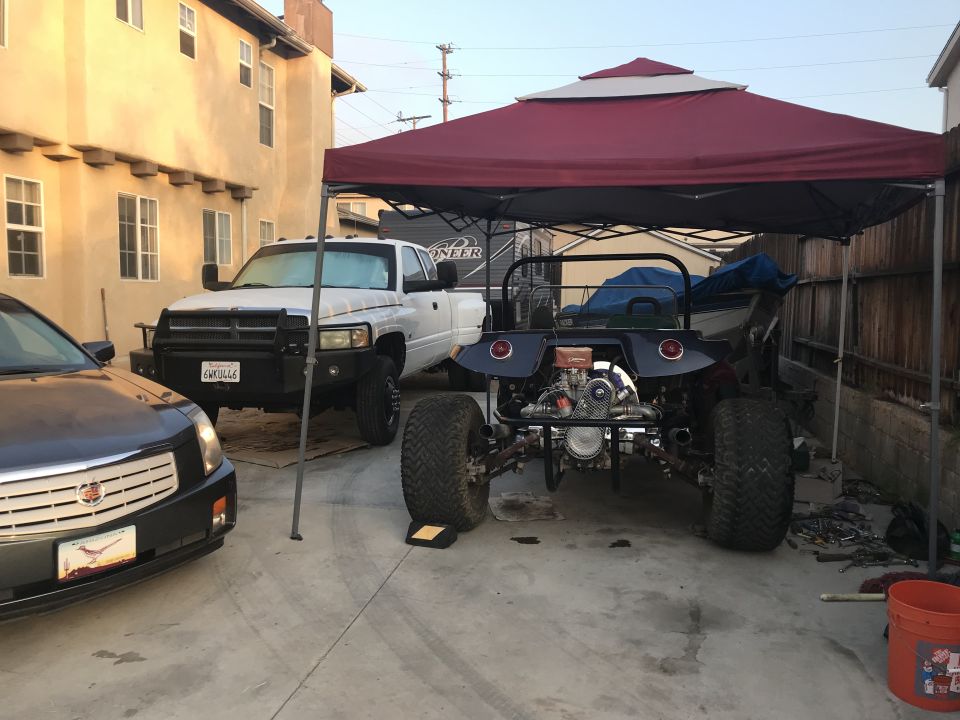 Custom dune buggy with exposed engine, parked under a canopy with a pickup truck and sedan.