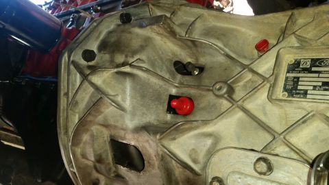 1985 Ford Bronco - A close-up view of a silver-colored transmission housing with two red caps and a metal data plate. The data plate has text that includes "Z…