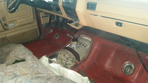 1985 Ford Bronco - The interior of a vehicle shows a removed floor carpet and exposed red floor pan. A manual transmission and shifter are visible in the cent…