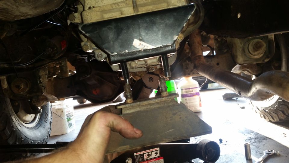Close-up of a hand holding a metal bracket under a vehicle's drivetrain.