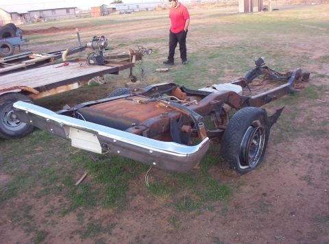 1988 Ford Bronco - A rusted vehicle frame sits on the ground, missing its body and engine, with a chrome bumper and a flat tire visible.