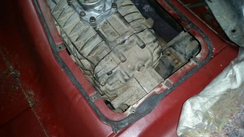 1985 Ford Bronco - A close-up view of a transfer case assembly mounted within the interior of a vehicle, surrounded by red carpeting and a black rubber seal.