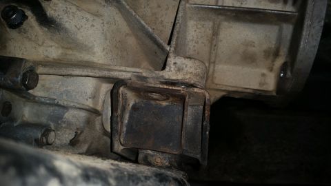 1985 Ford Bronco - A close-up, low-angle shot shows a rusty, square-shaped transmission mount bracket attached to the underside of a vehicle's drivetrain. The…
