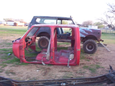 1988 Ford Bronco - Two vehicles, a red truck cab and a blue SUV, sit in a yard. The truck cab is missing its hood and fenders.
