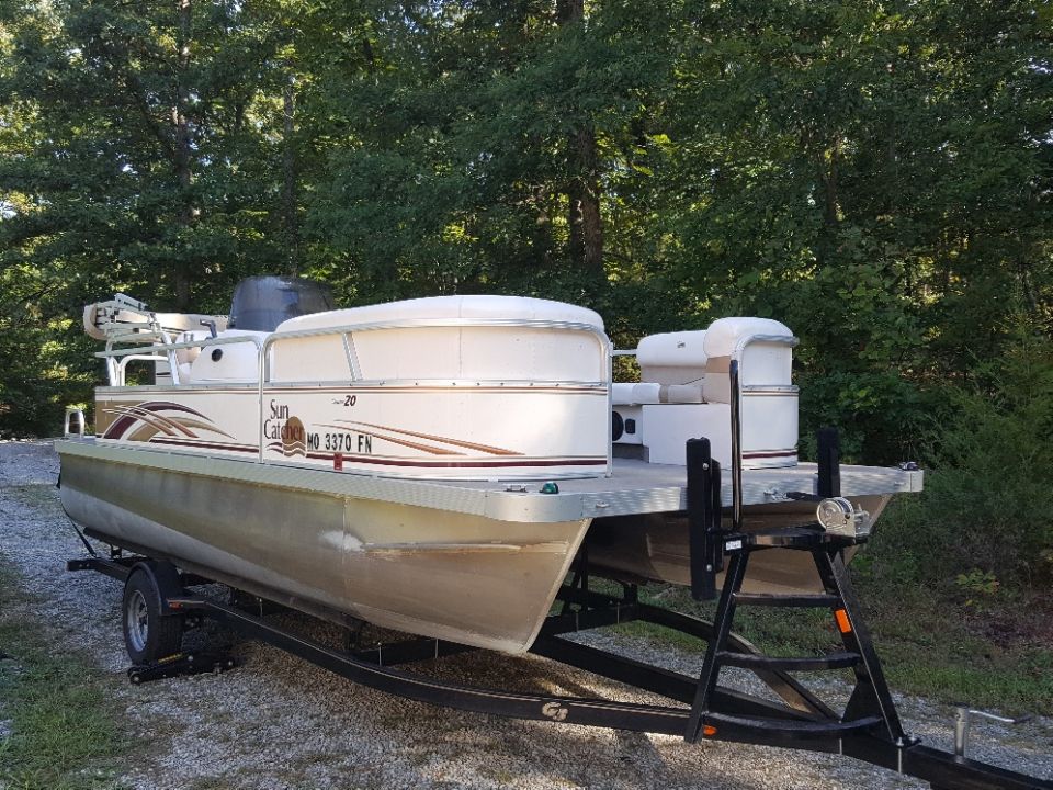 Beige and brown pontoon boat on a trailer in a wooded area.