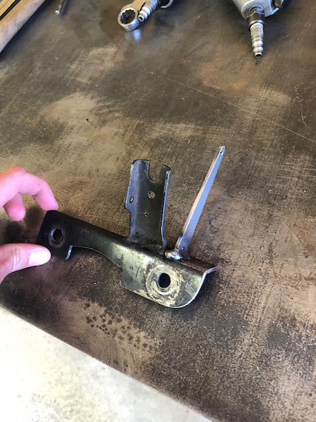 1981 Ford Bronco - A black metal bracket with two circular holes and a welded metal piece is held by a hand over a dark, textured workbench. Tools, including…