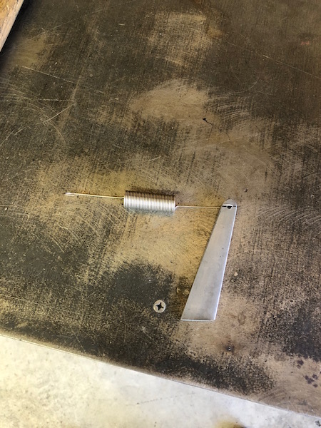 1981 Ford Bronco - A metal spring is attached to a small metal fin, which is screwed to a dark, textured surface. A single screw is visible near the fin.