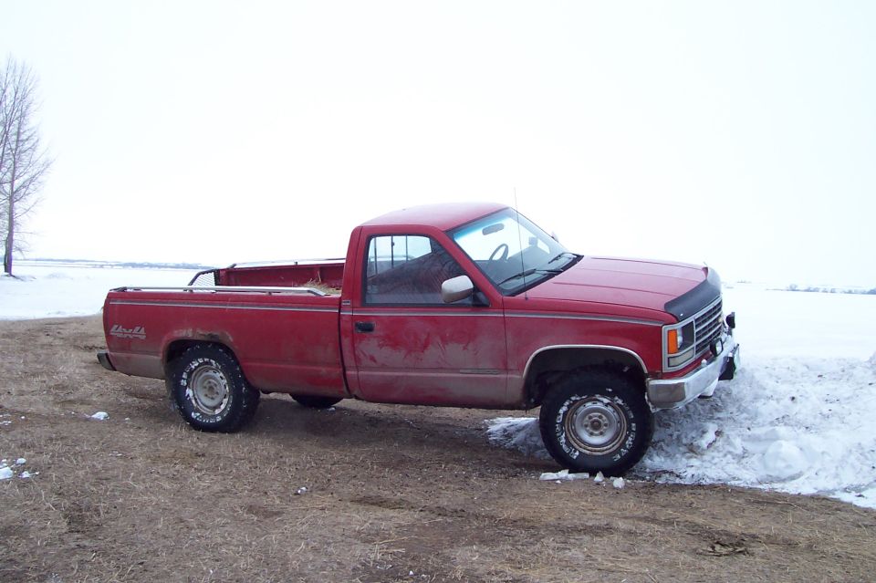 Red single-cab long-bed Chevy 4x4 pickup parked by a snowbank under gray sky