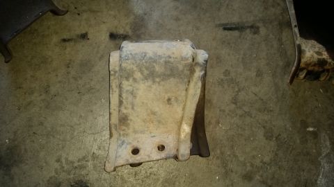 1985 Ford Bronco - A dirty, metal suspension part is shown lying on a concrete floor. The part has two circular holes at the bottom and appears to be a bracke…