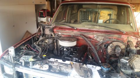 1985 Ford Bronco - The engine bay of a red Ford Bronco is shown with the hood open, revealing the engine, air filter, and various hoses and wires. Tools and a…