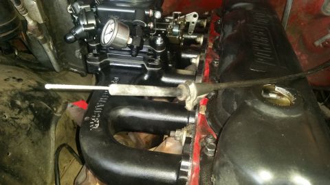 1985 Ford Bronco - A close-up view of a black carburetor and intake manifold on a red engine block. A throttle cable extends across the frame, and a fuel pres…