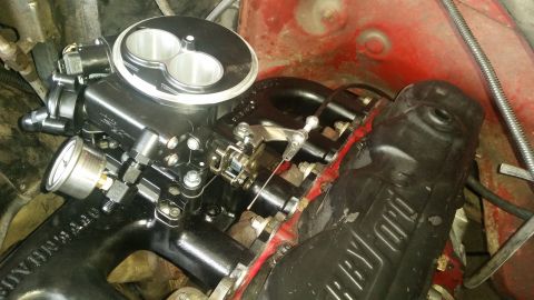1985 Ford Bronco - A close-up, high-angle view of a black aftermarket throttle body and intake manifold installed on a red engine bay. The throttle body has t…