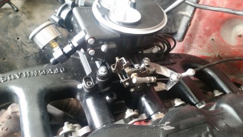 1985 Ford Bronco - A close-up view of a black aftermarket intake manifold with a carburetor and throttle linkage installed on a red vehicle engine. A fuel pre…