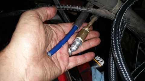 1985 Ford Bronco - A person's hand holds a blue hose and a brass fitting with a silver connector. Braided black hoses are visible in the background.