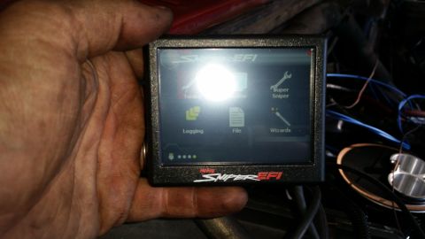 1985 Ford Bronco - A hand holds a Holley Sniper EFI handheld tuner with a menu displaying options like Logging, File, and Wizards. The device is black with a…
