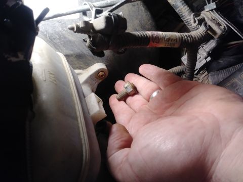 1996 Ford Bronco - A hand holds a bolt with a washer and nut in front of a white plastic reservoir and engine wiring harness.