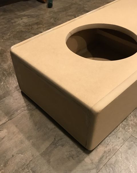 1995 Ford Bronco - A tan, unfinished speaker box with a circular cutout sits on a grey tiled floor.