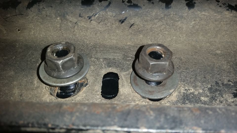 Two bolts with nuts and washers on a dirty metal bracket with a hole between them.