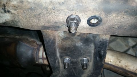 1985 Ford Bronco - A close-up view shows a bolt with a washer securing a metal bracket to the underside of a vehicle. Below the bracket, two nuts are visible…