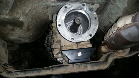 1985 Ford Bronco - The underside of a vehicle shows a transmission and transfer case assembly, with a catalytic converter visible to the right. A silver flang…