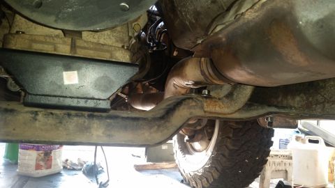 1985 Ford Bronco - Underside view of a vehicle's drivetrain and exhaust system, showing the transfer case, exhaust pipes, and a portion of the frame and tire.