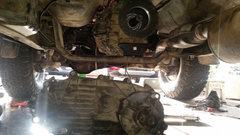 1985 Ford Bronco - The underside of a vehicle, likely a Ford Bronco, shows a transmission assembly being worked on. The transmission is detached and resting o…
