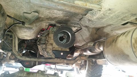 1985 Ford Bronco - The underside of a vehicle shows the transmission, exhaust system, and part of the drivetrain. A circular clutch housing is visible in the…
